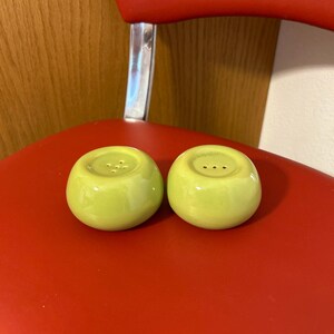 May include: Two light green ceramic salt and pepper shakers. Each shaker is round with a small hole pattern on top. The shakers are sitting on a red surface.