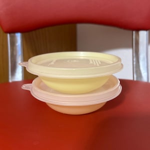 May include: Two yellow and peach-colored plastic bowls with lids are stacked on top of each other. The bowls have a rounded shape and are covered with light-colored lids. The bowls are sitting on a red surface.