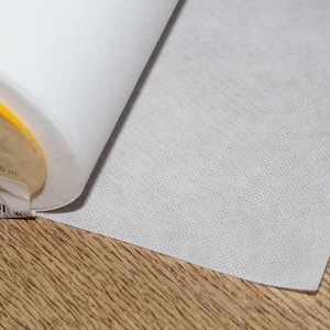 Puede incluir: Un rollo de tela blanca con una superficie texturizada, parcialmente desenrollado para revelar el material. La tela está sobre una superficie de madera. Una etiqueta con texto es visible en el rollo.