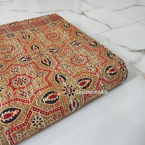 May include: A brown fabric with a red, black, and white geometric pattern. The fabric is folded in half and has a white stitching detail. The text "Textileofindia" is visible in the lower right corner of the image.