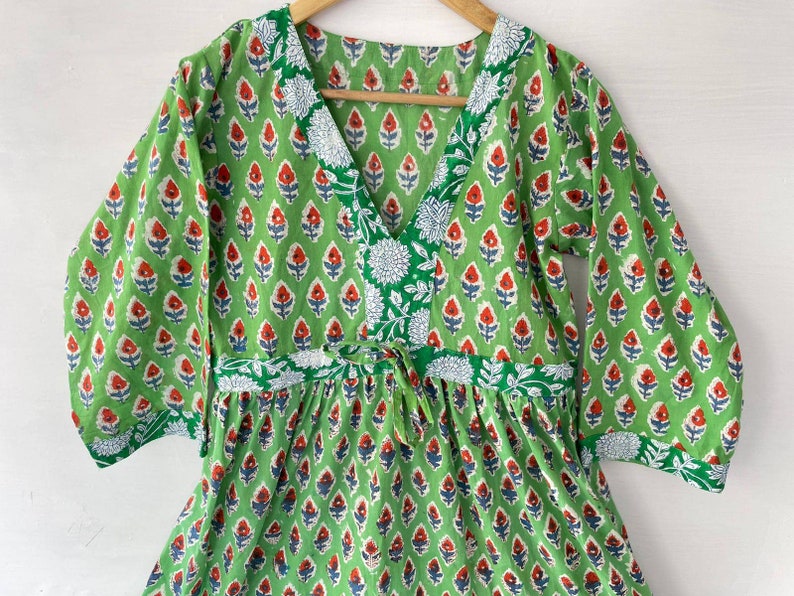 May include: A green cotton dress with a V-neck and three-quarter sleeves. The dress features a repeating floral pattern in red, white, and blue on a green background. The neckline and waist have white floral accents.