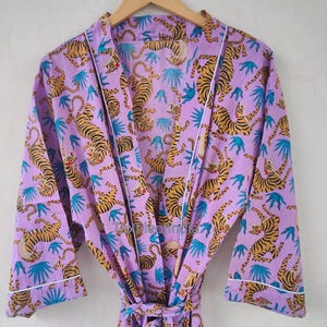 May include: A lilac-colored robe with a repeating pattern of orange tigers and blue leaf shapes. The robe has white trim along the edges and a matching belt. The robe is hanging on a wooden hanger.