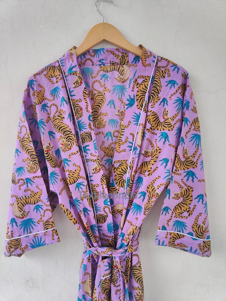May include: A lilac-colored robe with a repeating pattern of orange tigers and blue leaf shapes. The robe has white trim along the edges and a matching belt. The robe is hanging on a wooden hanger.