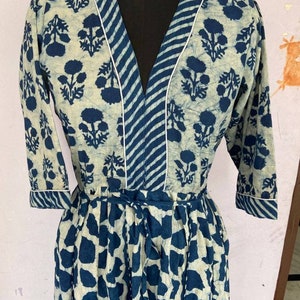 May include: A long-sleeved dress with a V-neck and a tie waist. The dress features a floral pattern in navy blue on a cream background. The neckline and cuffs are trimmed with white piping. The lower portion of the dress has a navy blue and cream pattern.