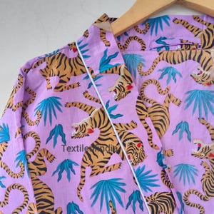 May include: A lilac-colored robe with a repeating pattern of tigers and teal-colored tropical leaves. The robe has white trim and is hanging on a wooden hanger. The text "Textile India" is visible on the robe.