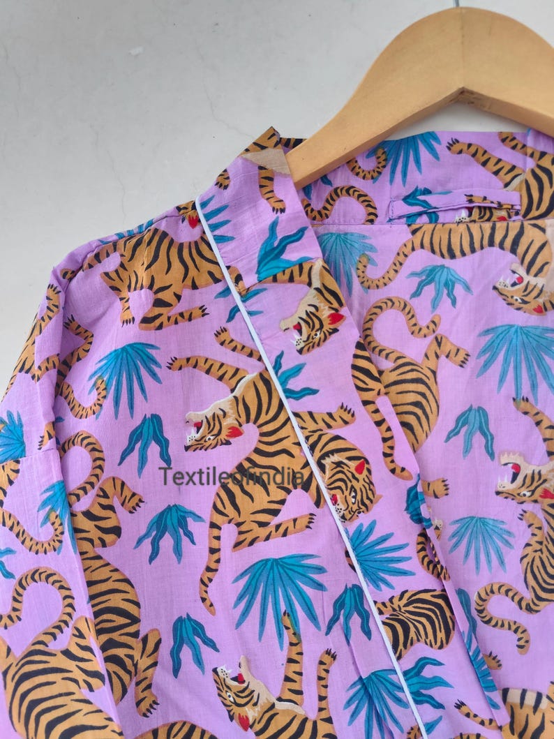 May include: A lilac-colored robe with a repeating pattern of tigers and teal-colored tropical leaves. The robe has white trim and is hanging on a wooden hanger. The text "Textile India" is visible on the robe.