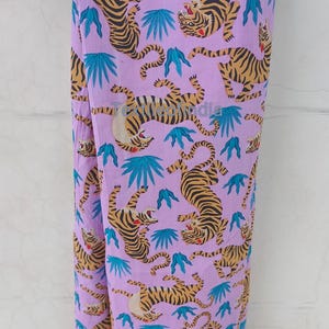 May include: A lavender fabric with a repeating pattern of tigers in shades of orange and black, interspersed with teal-colored leaf shapes. The tigers are depicted in various poses, creating a dynamic and eye-catching design.