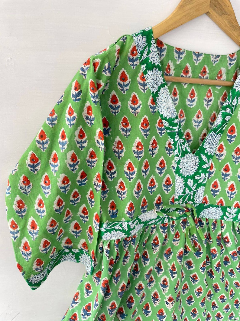 May include: A green, short-sleeved dress with a V-neck and a floral pattern. The pattern features red and blue flowers on a green background. The dress has a tie at the waist and white floral trim.