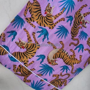May include: A lilac-colored fabric featuring a repeating pattern of orange and black tigers and teal-colored tropical leaves. The fabric is trimmed with a white border. The tigers are in various poses, some roaring.
