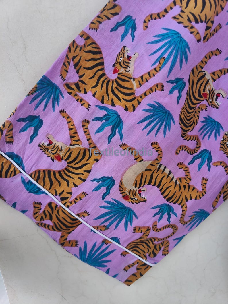 May include: A lilac-colored fabric featuring a repeating pattern of orange and black tigers and teal-colored tropical leaves. The fabric is trimmed with a white border. The tigers are in various poses, some roaring.