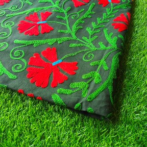May include: A green fabric tablecloth with red and yellow embroidered flowers. The tablecloth is folded and lying on a green grassy surface.
