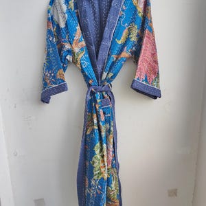 May include: A blue, floral-patterned robe with a matching belt, hanging on a wooden hanger. The robe features a patchwork design with various colors, including blue, pink, and white. The robe is made of a soft, quilted fabric.