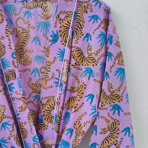 May include: A lilac-colored robe with a repeating pattern of tigers and blue tropical leaves. The tigers have orange and black stripes. The robe has white trim along the edges and is hanging on a wooden hanger.