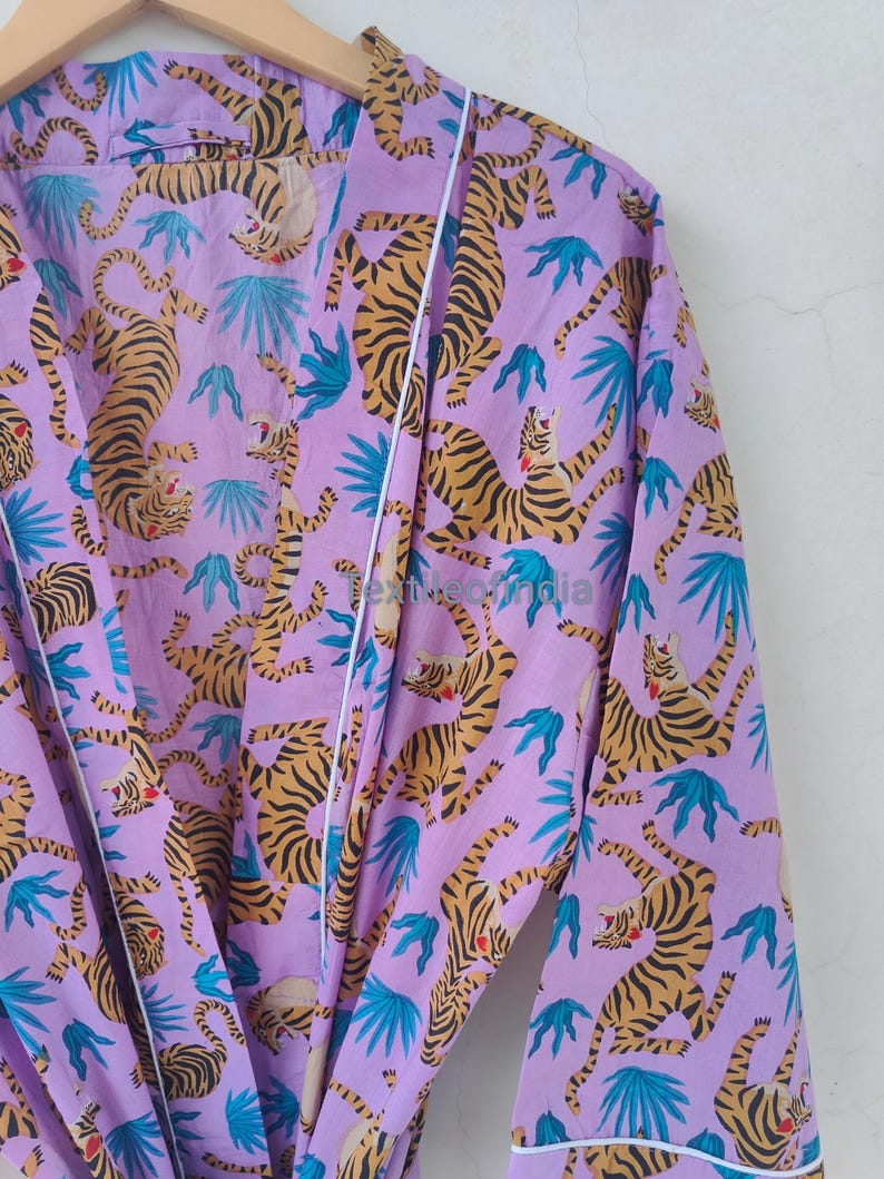 May include: A lilac-colored robe with a repeating pattern of tigers and blue tropical leaves. The tigers have orange and black stripes. The robe has white trim along the edges and is hanging on a wooden hanger.