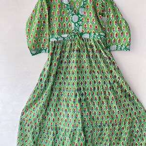 May include: A green dress with a V-neck and long sleeves, adorned with a repeating floral pattern in red and blue. The dress features a drawstring waist and is made of a lightweight fabric, perfect for warm weather. The dress is hanging on a wooden hanger.