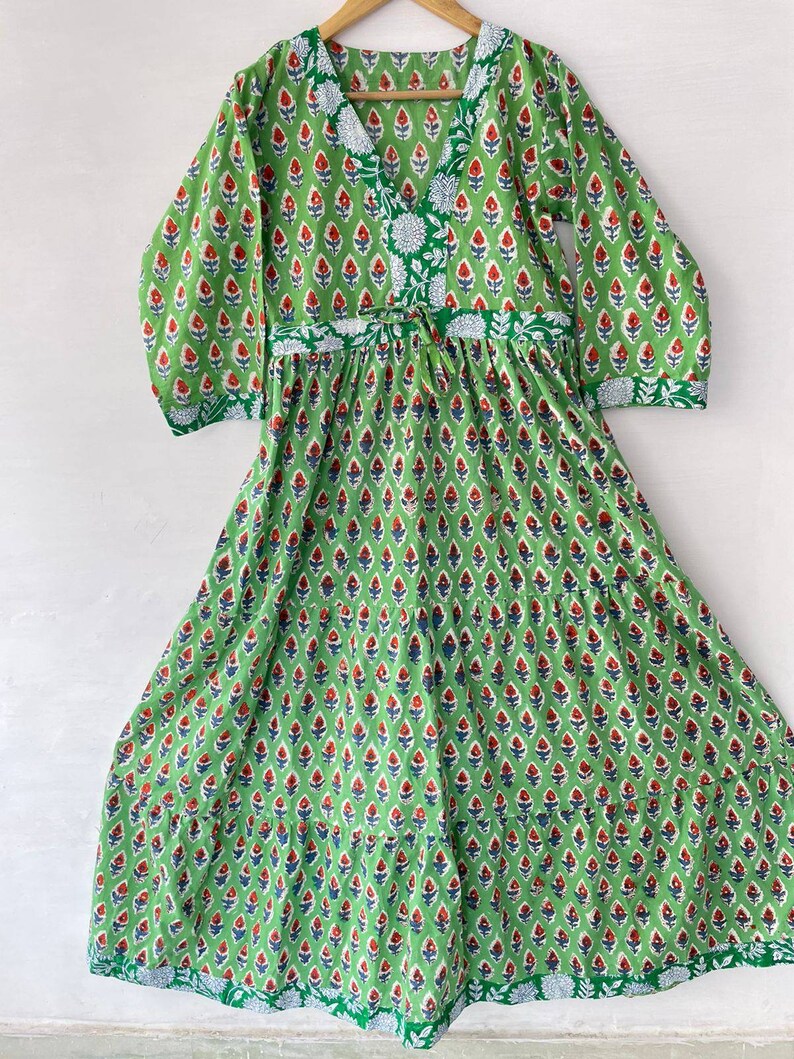May include: A green dress with a V-neck and long sleeves, adorned with a repeating floral pattern in red and blue. The dress features a drawstring waist and is made of a lightweight fabric, perfect for warm weather. The dress is hanging on a wooden hanger.