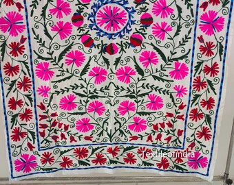 May include: White textile with a colourful floral embroidery design. The embroidery features pink, red, and blue flowers with green leaves. The design is symmetrical, with a central circular pattern and a border. The text "textileofindia" is visible.