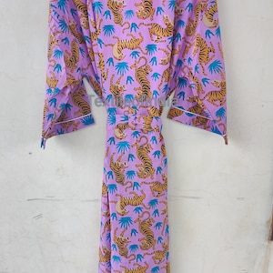 May include: A lilac-colored robe with a repeating pattern of tigers and blue leaf-like shapes. The robe has a white trim and a matching belt. The robe is hanging on a wooden hanger.