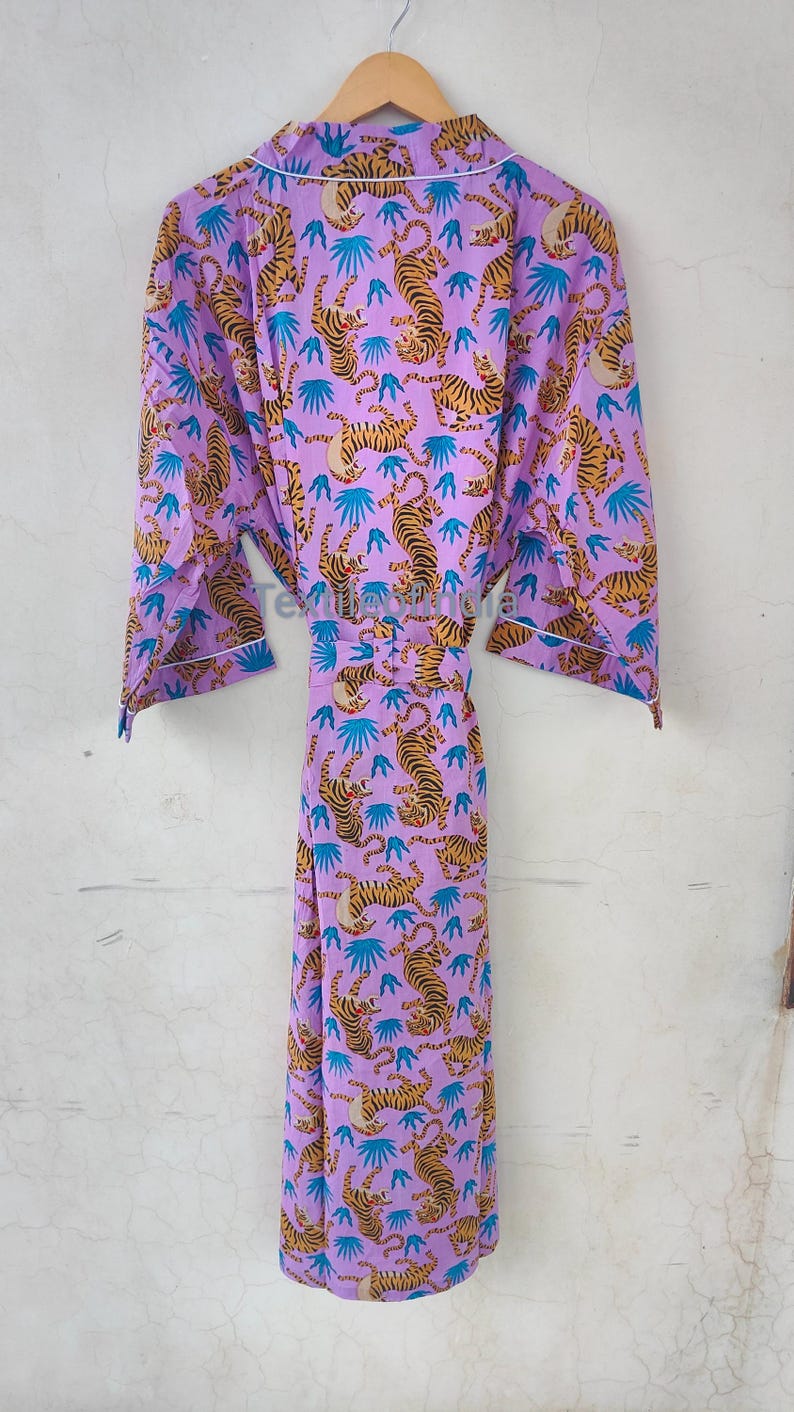 May include: A lilac-colored robe with a repeating pattern of tigers and blue leaf-like shapes. The robe has a white trim and a matching belt. The robe is hanging on a wooden hanger.