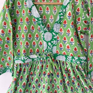May include: A green dress with a V-neck and long sleeves. The dress features a repeating floral pattern in red, white, and blue. The neckline and waist are accented with white floral designs.
