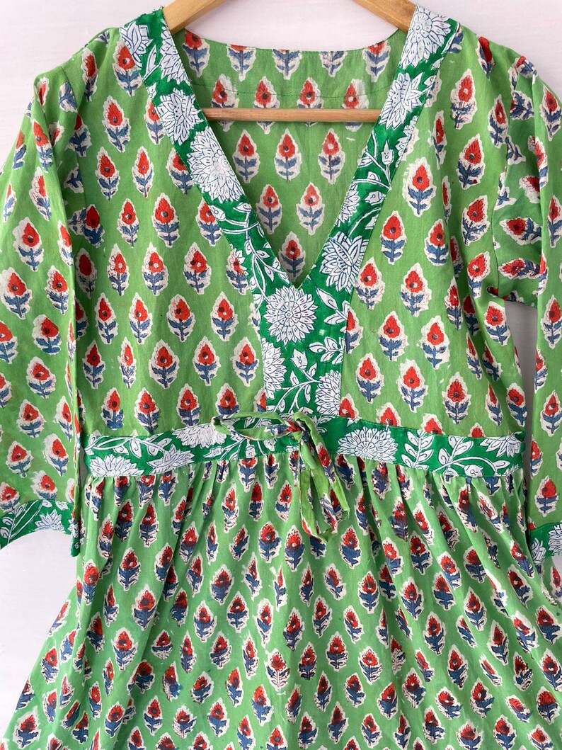 May include: A green dress with a V-neck and long sleeves. The dress features a repeating floral pattern in red, white, and blue. The neckline and waist are accented with white floral designs.
