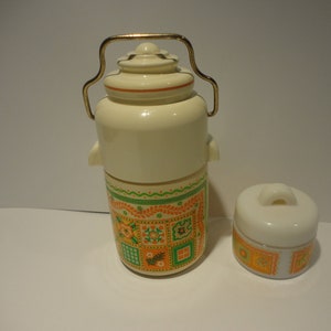以下が含まれることがあります： クリーム色のヴィンテージ魔法瓶と小さな容器。魔法瓶は金色のハンドルと、緑、オレンジ、黄色の花柄の装飾バンドが付いています。小さな容器にも同じデザインが施されています。