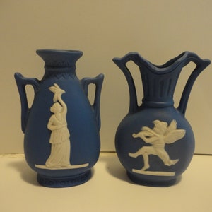 以下が含まれることがあります： 白で浮き彫りになったデザインが施された青と白のセラミック製の壺が2つ。1つの壺には鳥を持った女性が描かれ、もう1つの壺には楽器を演奏している羽根のある人物が描かれている。どちらの壺も青い背景に白いデザインが施されている。