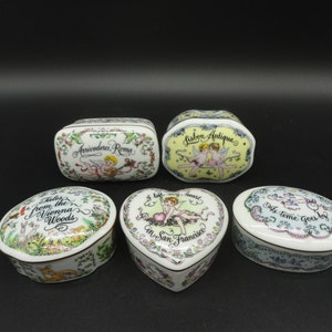 May include: Five porcelain trinket boxes with colorful floral designs and cherub figures. The boxes have different shapes, including oval, heart, and rectangular. The lids feature various inscriptions, including "Lisbon Antiqua", "Arrivederci, Roma", "Tales from the Vienna Woods", "I left my heart in San Francisco", and "As time goes by".