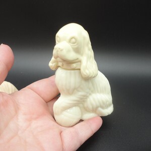 May include: Ivory-colored dog figurine with a detailed design. The dog is sitting upright, with its head turned slightly to the side. The figurine has a collar and a textured coat. The background is dark.