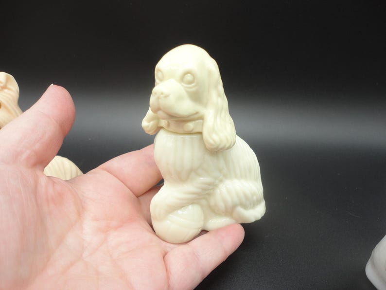 May include: Ivory-colored dog figurine with a detailed design. The dog is sitting upright, with its head turned slightly to the side. The figurine has a collar and a textured coat. The background is dark.