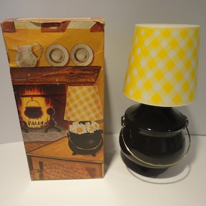 Puede incluir: Una lámpara negra con forma de caldero con una pantalla de lámpara a cuadros amarillos y blancos. La lámpara está sobre una mesa con una caja detrás. La caja tiene una imagen de una chimenea con un caldero colgando sobre el fuego.