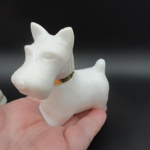 May include: A white figurine of a Scottish Terrier dog with a gold-colored collar. The dog is made of a smooth, opaque material and is posed in a standing position. The figurine is held in a person's hand against a black background.