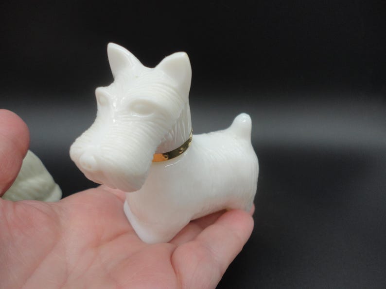 May include: A white figurine of a Scottish Terrier dog with a gold-colored collar. The dog is made of a smooth, opaque material and is posed in a standing position. The figurine is held in a person's hand against a black background.