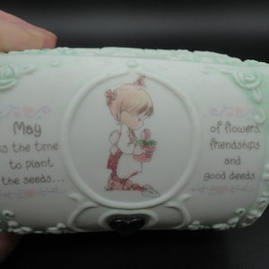 Puede incluir: Un joyero de cerámica blanca con un borde en relieve floral de color verde pálido. El centro presenta un niño de dibujos animados sosteniendo una flor en maceta. El texto dice: May is the time to plant the seeds... of flowers, friendships and good deeds. Un pequeño cierre negro en forma de corazón se encuentra en la parte inferior.