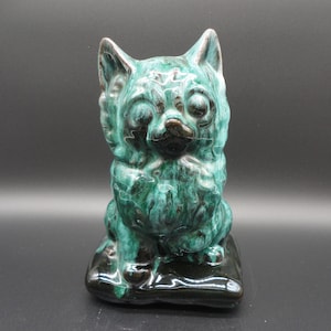 Puede incluir: Una figurita de cerámica de un gato verde y negro sentado en un cojín negro. El gato tiene un aspecto esponjoso y mira hacia adelante.