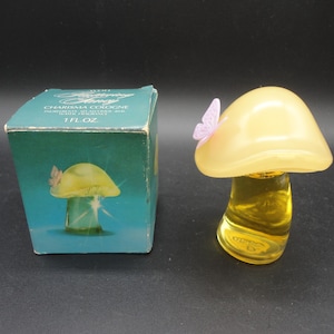 May include: A vintage perfume bottle shaped like a mushroom with a light yellow cap and a clear base filled with yellow liquid. A small purple butterfly adorns the cap. The box next to it is teal with a mushroom and butterfly illustration.