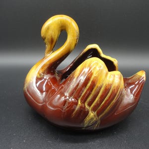 以下が含まれることがあります： 茶色と黄色の釉薬をかけたセラミック製の白鳥のプランター。白鳥は長い首と幅広い体を持っています。プランターは、小さな植物や花を飾るのに最適です。