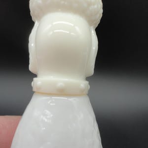 May include: A close-up of a white poodle figurine. The figurine has a textured body and a detailed head with a fluffy top. It wears a collar with small, round studs. The background is dark.