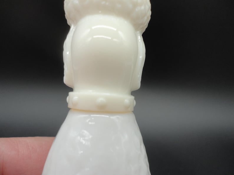 May include: A close-up of a white poodle figurine. The figurine has a textured body and a detailed head with a fluffy top. It wears a collar with small, round studs. The background is dark.