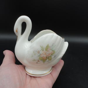 Puede incluir: Una figurita de cisne de porcelana blanca con ribetes dorados y un diseño floral. El cisne tiene un aspecto delicado y femenino y es perfecto para añadir un toque de elegancia a cualquier hogar.