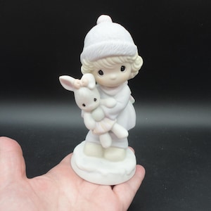 Puede incluir: Figura de porcelana de un niño con gorro y abrigo de invierno blancos, sosteniendo un conejito blanco con un lazo rosa. La figura está sobre una base blanca y tiene una expresión dulce e inocente. La paleta de colores es pastel.
