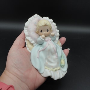 Puede incluir: Una figura de porcelana de un bebé con cabello rubio, vestido con una túnica azul claro y blanca con detalles en rosa. El bebé descansa sobre una almohada con volantes y un pequeño patrón floral azul. Un sonajero verde cuelga del vestido.