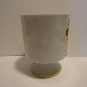 May include: A white ceramic mug with a yellow and black design. The mug has a footed base.