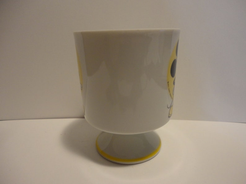 May include: A white ceramic mug with a yellow and black design. The mug has a footed base.