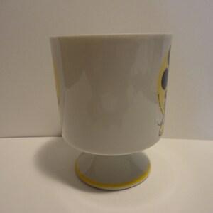 May include: A white ceramic mug with a yellow and black design. The mug has a footed base.