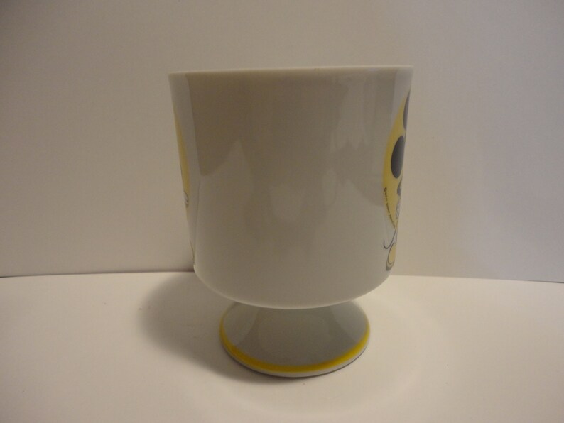 May include: A white ceramic mug with a yellow and black design. The mug has a footed base.