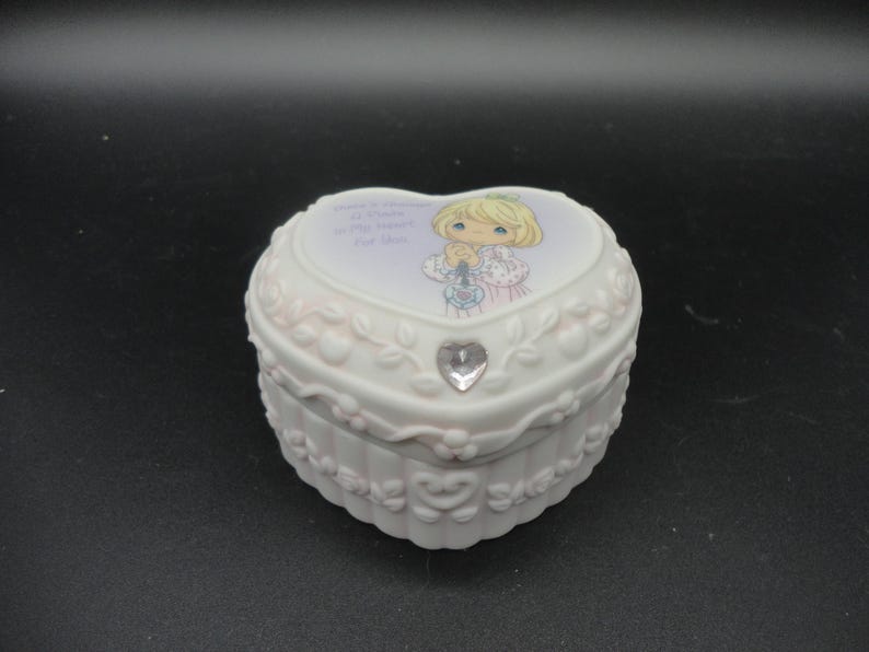 Puede incluir: Un joyero de porcelana blanca en forma de coraz&oacute;n con un relieve floral y un cristal transparente. La tapa muestra una ilustraci&oacute;n de un ni&ntilde;o rubio con las manos juntas y el texto There is Always a Place in My Heart for You.