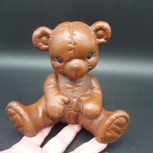 Puede incluir: Una figurita de oso de peluche marrón con un diseño cosido. El oso está sentado con las piernas cruzadas y tiene una expresión dulce en su rostro.