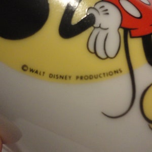 May include: A white ceramic plate featuring a cartoon image of Mickey Mouse. The plate has a yellow background and depicts Mickey Mouse in black and red, with a black tail. The plate is marked with the copyright text "&copy; WALT DISNEY PRODUCTIONS".