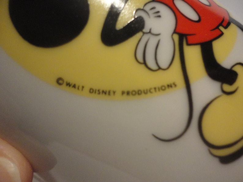 May include: A white ceramic plate featuring a cartoon image of Mickey Mouse. The plate has a yellow background and depicts Mickey Mouse in black and red, with a black tail. The plate is marked with the copyright text "&copy; WALT DISNEY PRODUCTIONS".